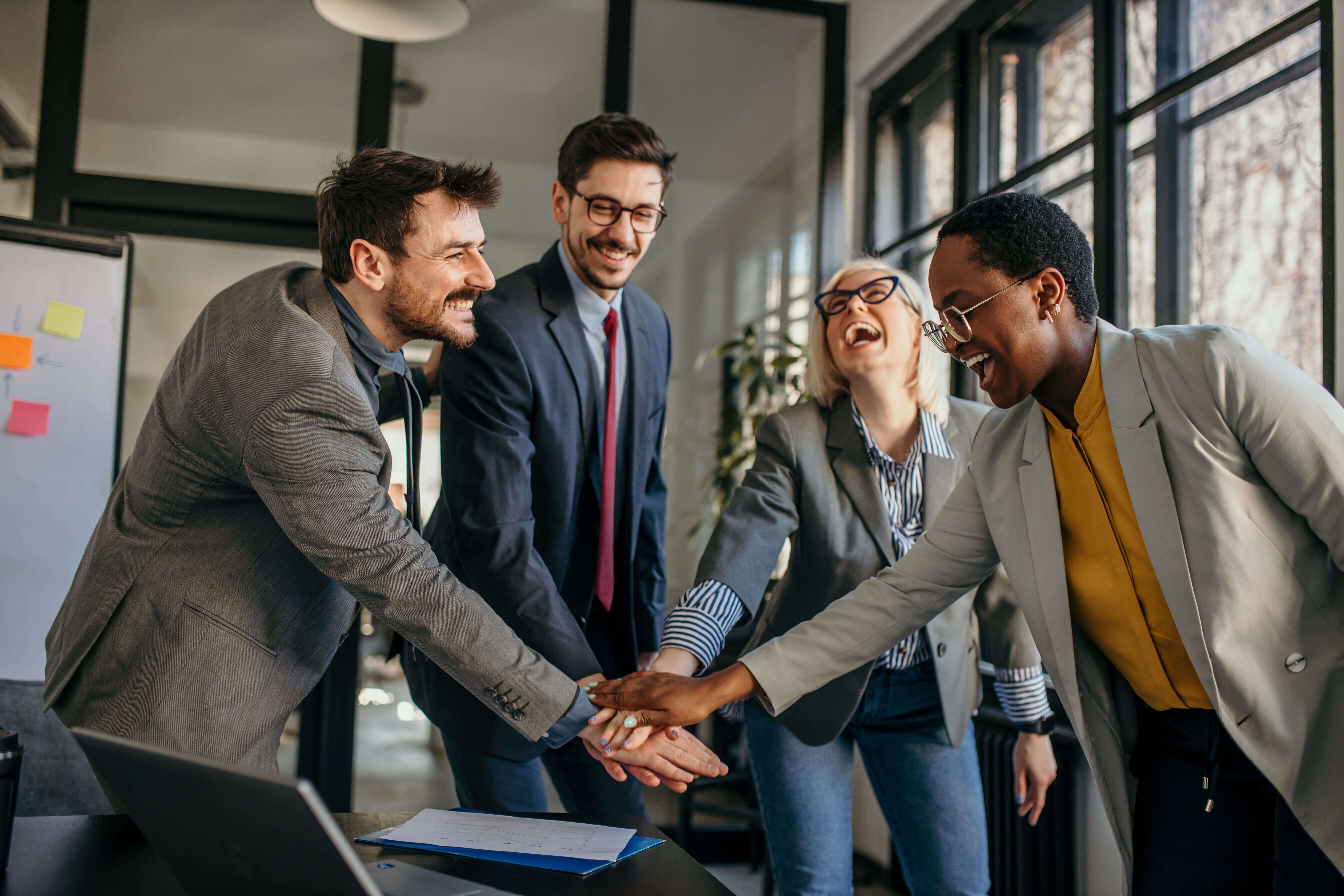 Four colleagues smiling and shaking hands in a bright office setting.